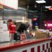Close-up of a restaurant counter and food-service area, with a blurred employee in the background and distant emergency lights visible outside.