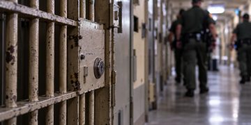 Close-up of a secured California prison corridor with a barred cell door and blurred correctional officers in the background, illustrating a homicide investigation inside Pelican Bay State Prison.