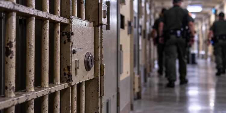 Close-up of a secured California prison corridor with a barred cell door and blurred correctional officers in the background, illustrating a homicide investigation inside Pelican Bay State Prison.