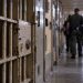 Close-up of a secured California prison corridor with a barred cell door and blurred correctional officers in the background, illustrating a homicide investigation inside Pelican Bay State Prison.