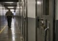 Close-up of a secured prison corridor and metal door, with a blurred correctional employee walking in the background.