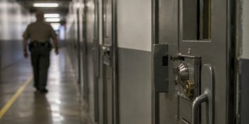 Close-up of a secured prison corridor and metal door, with a blurred correctional employee walking in the background.
