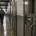 Close-up of a secured prison corridor and metal door, with a blurred correctional employee walking in the background.