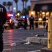 Close-up of a security guard in a parking lot with a blurred bar entrance, police vehicles and emergency responders in the background.
