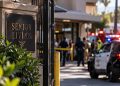 Close-up of a senior living facility entrance with blurred police vehicles and emergency responders outside after a workplace shooting in South Los Angeles.
