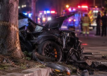 Close-up of a severely damaged sports car near a roadside tree at night, with blurred emergency lights and responders in the background, illustrating a fatal crash in Irvine.