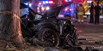 Close-up of a severely damaged sports car near a roadside tree at night, with blurred emergency lights and responders in the background, illustrating a fatal crash in Irvine.