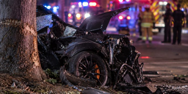 Close-up of a severely damaged sports car near a roadside tree at night, with blurred emergency lights and responders in the background, illustrating a fatal crash in Irvine.