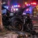 Close-up of a severely damaged sports car near a roadside tree at night, with blurred emergency lights and responders in the background, illustrating a fatal crash in Irvine.