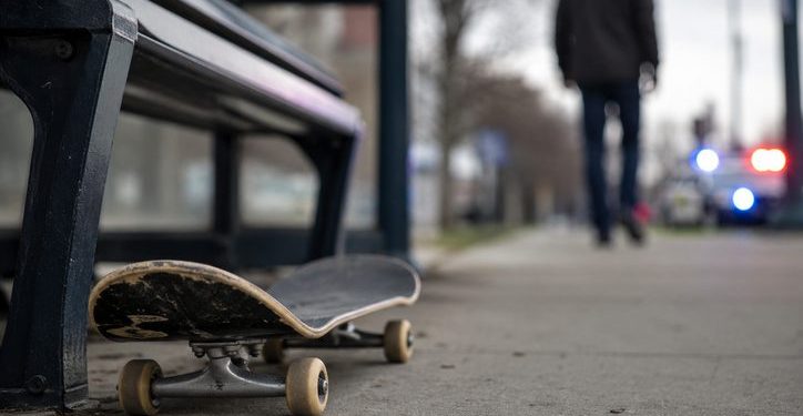 Close-up of a skateboard lying beside a bus stop bench, with a blurred pedestrian and distant police presence visible in the background.