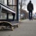 Close-up of a skateboard lying beside a bus stop bench, with a blurred pedestrian and distant police presence visible in the background.
