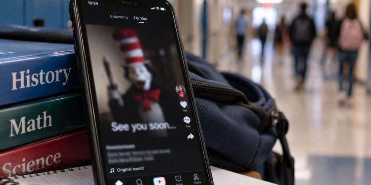 Close-up of a smartphone displaying a blurred social media post beside school supplies, with a school hallway softly illustrating a viral online trend targeting school communities.