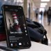 Close-up of a smartphone displaying a blurred social media post beside school supplies, with a school hallway softly illustrating a viral online trend targeting school communities.