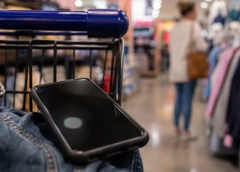 Close-up of a smartphone near a shopping cart inside a retail store, with a blurred shopper and store aisle visible in the background.