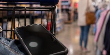 Close-up of a smartphone near a shopping cart inside a retail store, with a blurred shopper and store aisle visible in the background.