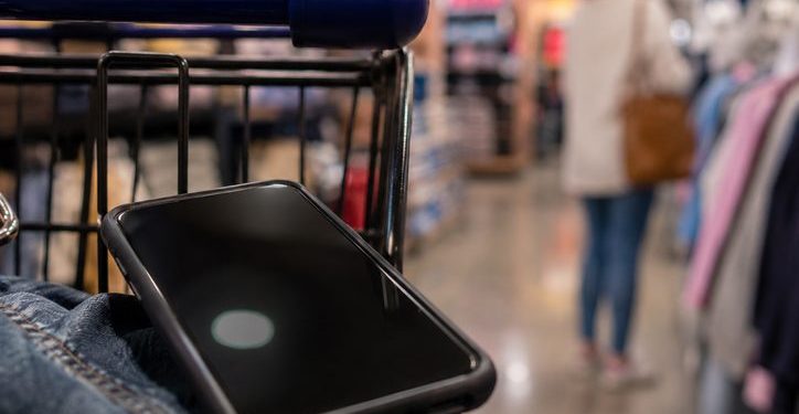 Close-up of a smartphone near a shopping cart inside a retail store, with a blurred shopper and store aisle visible in the background.