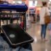 Close-up of a smartphone near a shopping cart inside a retail store, with a blurred shopper and store aisle visible in the background.