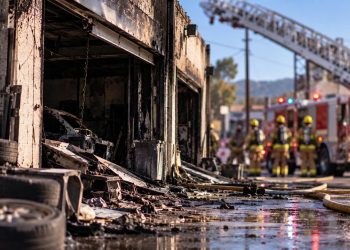 Close-up of a smoke-damaged auto body shop with firefighters and a ladder truck blurred in the background on a Newhall commercial street.
