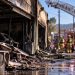 Close-up of a smoke-damaged auto body shop with firefighters and a ladder truck blurred in the background on a Newhall commercial street.