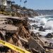 Close-up of a sun-baked Southern California coastline with large waves and damaged shoreline structures in the background, illustrating extreme heat and coastal recovery.