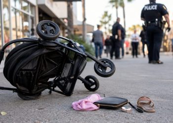 Close-up of a tipped stroller near a shopping center walkway, with blurred officers and pedestrians visible in the background.