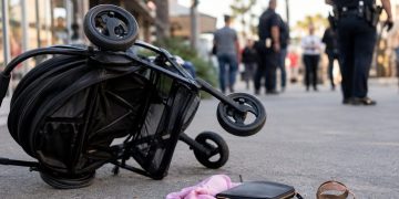Close-up of a tipped stroller near a shopping center walkway, with blurred officers and pedestrians visible in the background.