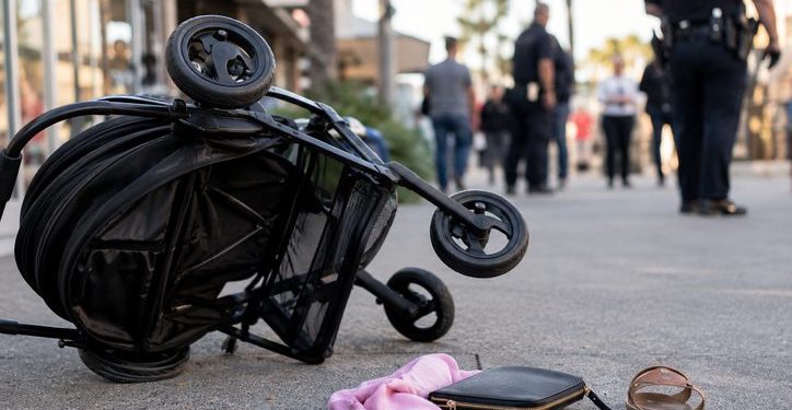 Close-up of a tipped stroller near a shopping center walkway, with blurred officers and pedestrians visible in the background.
