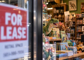 Close-up of a toy and book store window displaying dinosaur toys, children's books and stuffed animals, with a blurred “FOR LEASE” sign in the foreground.