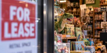 Close-up of a toy and book store window displaying dinosaur toys, children's books and stuffed animals, with a blurred “FOR LEASE” sign in the foreground.