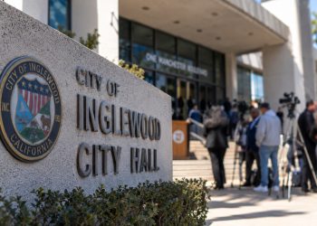 Close-up of an Inglewood City Hall exterior with blurred community members and officials in the background, illustrating scrutiny of the mayor.