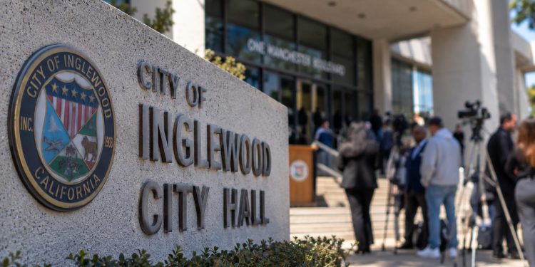 Close-up of an Inglewood City Hall exterior with blurred community members and officials in the background, illustrating scrutiny of the mayor.