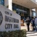 Close-up of an Inglewood City Hall exterior with blurred community members and officials in the background, illustrating scrutiny of the mayor.