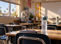Close-up of an empty classroom with a water bottle, portable fan and desks in warm sunlight, illustrating heat challenges at Southern California schools.