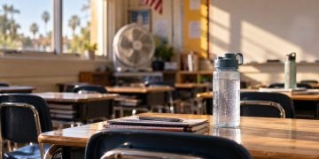Close-up of an empty classroom with a water bottle, portable fan and desks in warm sunlight, illustrating heat challenges at Southern California schools.
