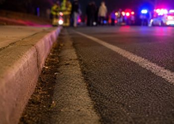 Close-up of an empty nighttime roadway near a curb, with blurred emergency lights and several responders standing in the distance.