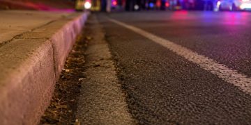 Close-up of an empty nighttime roadway near a curb, with blurred emergency lights and several responders standing in the distance.