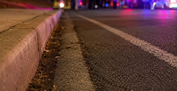 Close-up of an empty nighttime roadway near a curb, with blurred emergency lights and several responders standing in the distance.