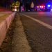 Close-up of an empty nighttime roadway near a curb, with blurred emergency lights and several responders standing in the distance.