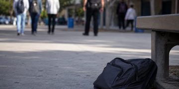 Close-up of an empty school walkway and backpack near a campus bench, with blurred students and a school safety officer visible in the distance.
