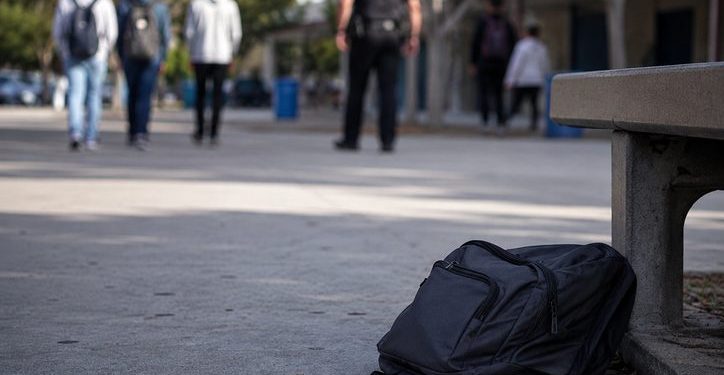 Close-up of an empty school walkway and backpack near a campus bench, with blurred students and a school safety officer visible in the distance.