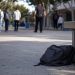 Close-up of an empty school walkway and backpack near a campus bench, with blurred students and a school safety officer visible in the distance.