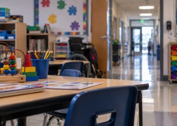 Close-up of an empty special education classroom with small desks and learning materials, with a blurred elementary school hallway in the background.