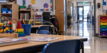 Close-up of an empty special education classroom with small desks and learning materials, with a blurred elementary school hallway in the background.