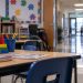 Close-up of an empty special education classroom with small desks and learning materials, with a blurred elementary school hallway in the background.
