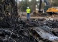 Close-up of charred debris and scorched fencing at an abandoned lot, with a blurred cleanup worker and heavy equipment in the background.