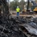 Close-up of charred debris and scorched fencing at an abandoned lot, with a blurred cleanup worker and heavy equipment in the background.