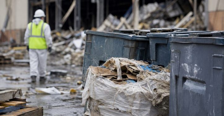 Close-up of cleanup debris and industrial waste containers outside a damaged warehouse, with a blurred cleanup worker in protective gear visible in the background.