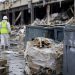 Close-up of cleanup debris and industrial waste containers outside a damaged warehouse, with a blurred cleanup worker in protective gear visible in the background.