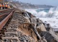 Close-up of coastal railroad tracks beside eroded sand and protective rocks, with large waves and a blurred shoreline in the background, illustrating rail repairs in San Clemente.