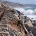 Close-up of coastal railroad tracks beside eroded sand and protective rocks, with large waves and a blurred shoreline in the background, illustrating rail repairs in San Clemente.